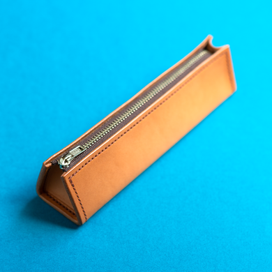 Full-grain hand-stitched brown leather pencil case with a zipper on a blue background