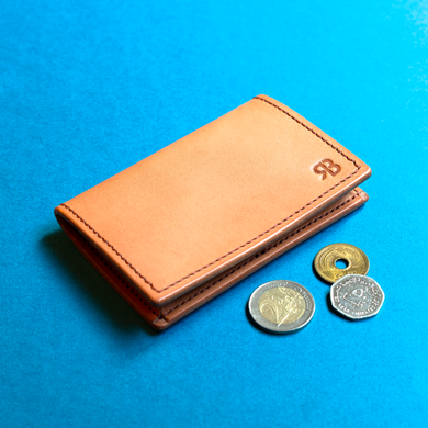Full-grain hand-stitched brown leather coin purse with coins on a blue background, closed