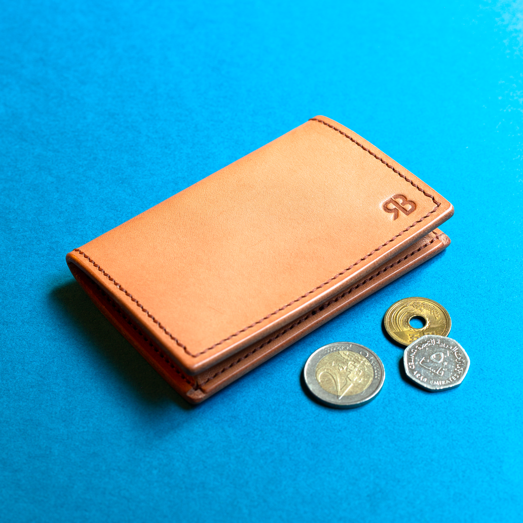 Full-grain hand-stitched brown leather coin purse with coins on a blue background, closed